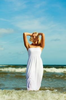 Attractive blonde woman wearing long white romantic dress walking in water sea during summer.. Blonde woman wearing dress walking in water