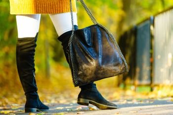 Woman presenting black suede high knee black boots holding big hand bag. Autumnal fashion, autumn season styled outfits. Female having a walk in park. Woman presenting high knee black boots
