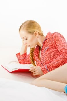 Girl lying in bed reading book. Young blonde female wearing red dotted pajamas relaxing at home on mattress.. Woman relaxing in bed reading book
