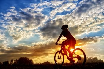 Silhouette of a girl riding a bicycle on the background of the setting sun. Summer landscape. The concept of freedom, travel and healthy lifestyle. Silhouette of girl riding bicycle on background of setting sun