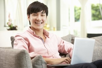 Young Chinese Man Using Laptop Whilst Relaxing On Sofa At Home