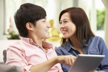 Young Chinese Couple Using Digital Tablet Whilst Relaxing On Sofa At Home