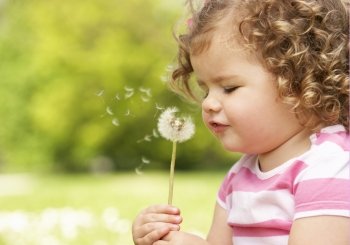 Young Girl In Summer Dress Sitting In Field Blowing Dandelion