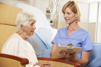 Nurse Taking Notes From Senior Female Patient Seated In Chair By Hospital Bed