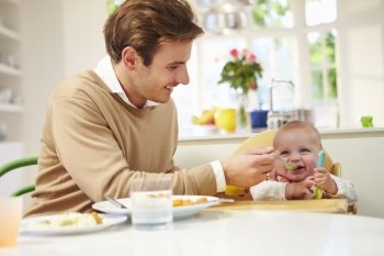 Father Feeding Baby Sitting In High Chair At Mealtime