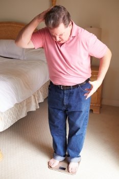 Overweight Man Weighing Himself On Scales In Bedroom