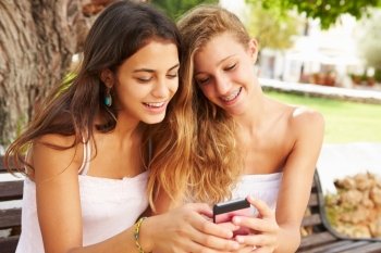 Two Teenage Girls Using Mobile Phone Sitting On Park Bench