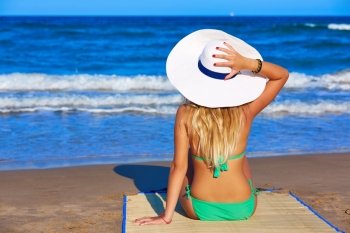 Blond young girl sitting sitting looking sea with beach hat rear back view