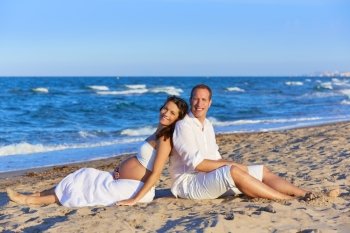 Beautiful couple pregnant woman in the beach sitting on sand happy together back to back