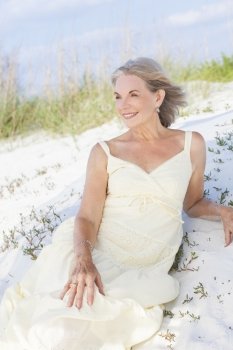 An attractive elegant classy senior woman in a yellow sun dress sitting on a white sand beach with grass and a blue sky behind her.