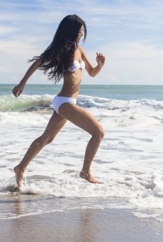 A sexy young brunette Asian woman or girl wearing a white bikini running on a deserted tropical beach with a blue sky 