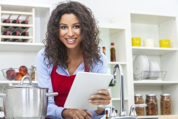 A beautiful happy young woman or girl wearing a red apron & using a tablet computer while cooking in her kitchen at home