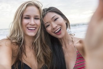 Two young women girls, one Asian Chinese, one blond, wearing bikinis on a beach, laughing taking selfie photograph with digital camera