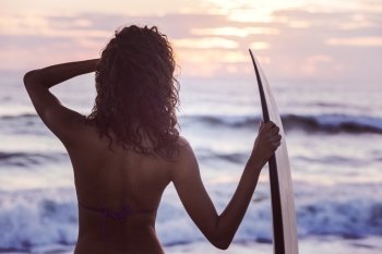 Instagram filter style rear view of beautiful sexy young woman surfer girl in bikini with surfboard on a beach at sunset or sunrise