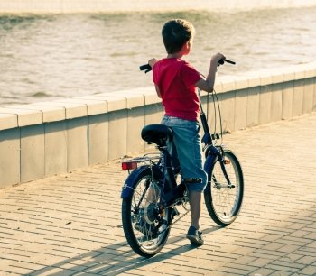 Backside view of a boy on bike near water. Red shirt, jeans shorts