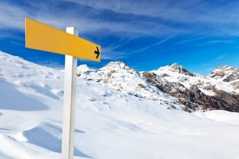 Yellow mountain guidepost along an alpine pathway over a snowy landscape in a clear winter day. Italian Alps, Val d’Aosta, Italy.