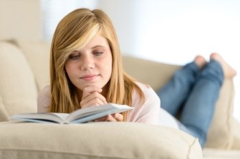 Young student girl reading book lying on sofa