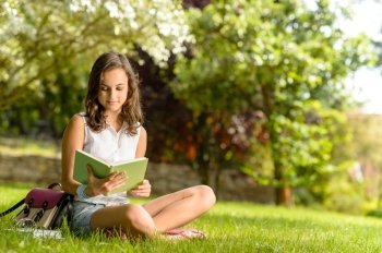 Student girl reading book sitting on grass in summer park
