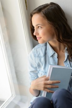 Beautiful teenage girl student holding book looking out the window