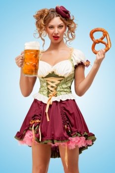 Young sexy Oktoberfest waitress wearing a traditional Bavarian dress dirndl holding a pretzel and beer mug, and pulling grimaces on blue background.