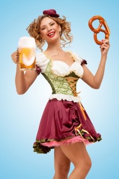 Young sexy Oktoberfest waitress wearing a traditional Bavarian dress dirndl holding a pretzel and beer mug, and laughing happily on blue background.
