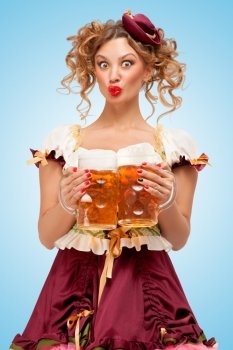 Young sexy Oktoberfest woman wearing a traditional Bavarian dress dirndl serving two beer mugs with happy smile on blue background.
