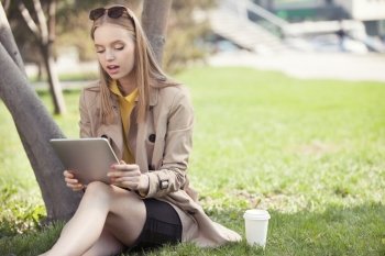 Beautiful young woman sitting on the grass with a disposable coffee cup, holding tablet in her hands, reading and studying against summer park background.
