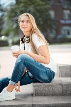 Happy young woman wearing vintage music headphones around her neck and sitting on stairs with a take away coffee cup against urban city background.