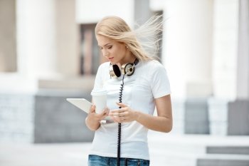 Happy young woman with vintage music headphones around her neck, holding a take away coffee cup and surfing internet on a tablet pc against urban city background.