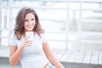 Beautiful young woman holding a take away coffee cup and sitting on the bridge against water background.