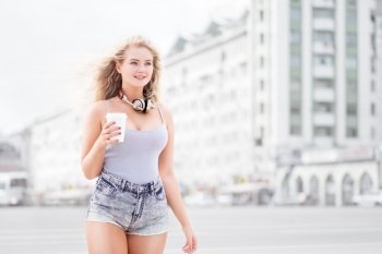 Happy young woman with music headphones, holding a take away coffee cup, listening to the music and walking against city background.