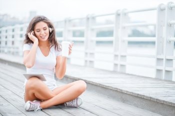 Happy young woman with music headphones and a take away coffee cup, surfing internet on tablet pc, listening to the music and sitting on the bridge.