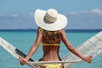 Woman in hammock on beach . Rear view of a woman in bikini with a stylish hat sitting on a hammock on a beach by the sea