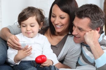 Toddler holding mobile telephone whilst sat with parents on sofa