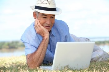 Senior man lying on the grass in the sunshine using a laptop computer