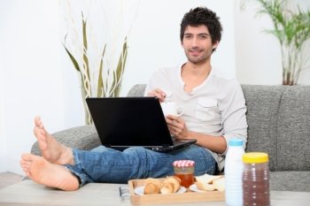 young man seated on a sofa with legs stretched out, doing computer and eating breakfast,