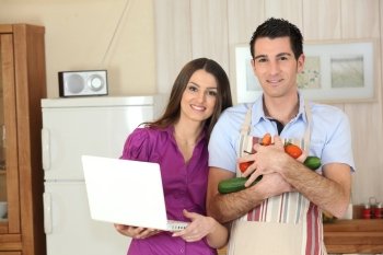 Couple in a kitchen with a laptop computer and arms full of vegetables