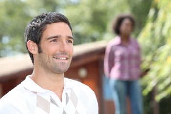 Man standing outside a log cabin with a woman out of focus in the background