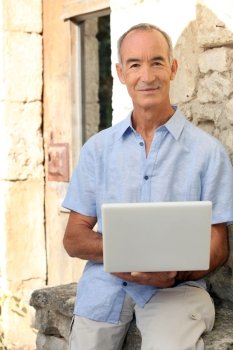 Grey haired man sat on stone wall with laptop