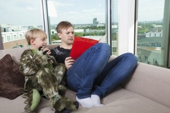 Boy dressed in dinosaur costume sitting with father reading story book on sofa bed at home
