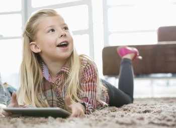Cute girl with digital tablet looking away while lying on rug in living room