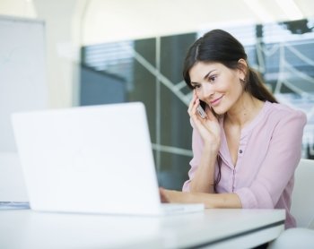 Beautiful young businesswoman using mobile phone while looking at laptop in office