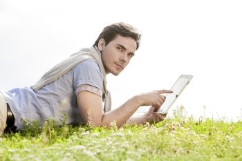 Side view of young man using digital tablet while lying on grass against clear sky