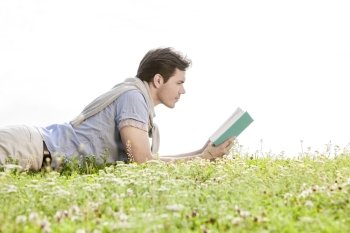 Side view of young man reading book while lying on grass against clear sky