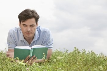 Young man reading book while lying on grass against sky