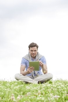 Full length of young man using digital tablet while sitting on grass against sky