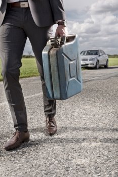 Low section of young businessman carrying gas can with broken car in background at countryside