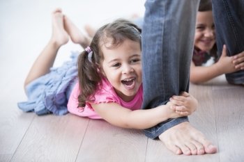 Playful girls holding father’s legs on hardwood floor