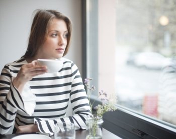 Thoughtful young woman looking out from window while drinking coffee in cafe