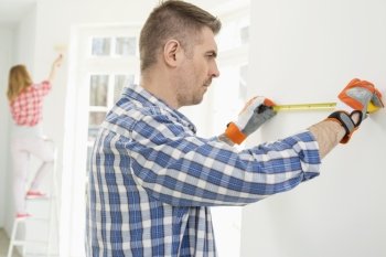 Man measuring wall with woman painting in background